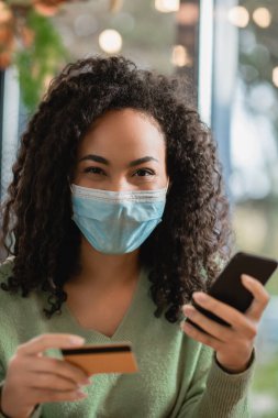 Kafede elinde akıllı telefon ve kredi kartı olan tıbbi maskeli Afro-Amerikan kadın.