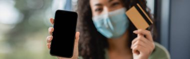 Beyaz ekranlı akıllı telefon ve bulanık arka planda tıbbi maskeli Afrikalı Amerikalı bir kadının elinde kredi kartı.