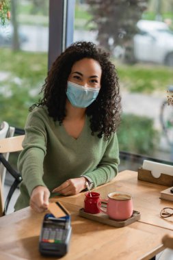 Kafe 'deki ödeme terminalinin yakınında sağlık maskeli Afro-Amerikan bir kadın elinde kredi kartı tutuyor. 