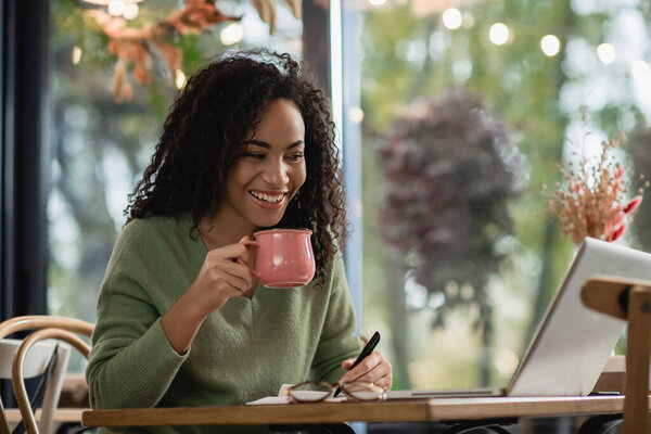 happy african american woman holding pen and cup of coffee while looking at laptop 