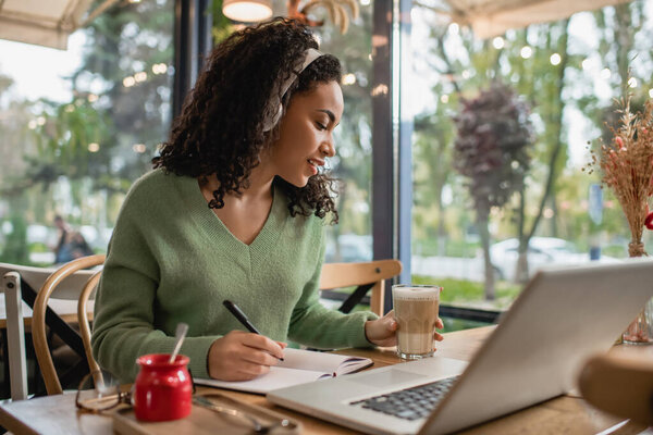 african american woman listening podcast in headphones and holding glass with coffee near laptop in cafe 