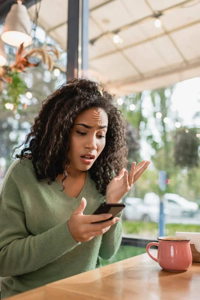 不满意的非洲裔美国女人一边在咖啡店摆姿势一边看智能手机 — 图库照片