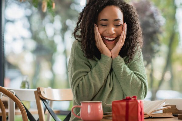 изумленная африканская американка смотрит на красную рождественскую подарочную коробку возле чашки в кафе