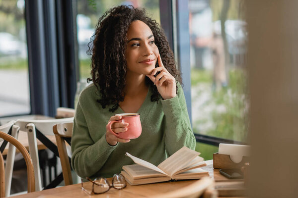 Мечтательная африканская американка держит чашку кофе и смотрит рядом с книгой на столе 