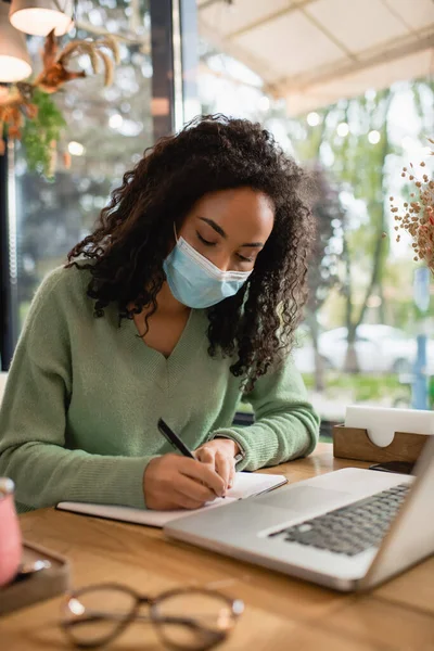 Mujer Afroamericana Escritura Máscara Médica Cuaderno Cerca Del Ordenador Portátil — Foto de Stock