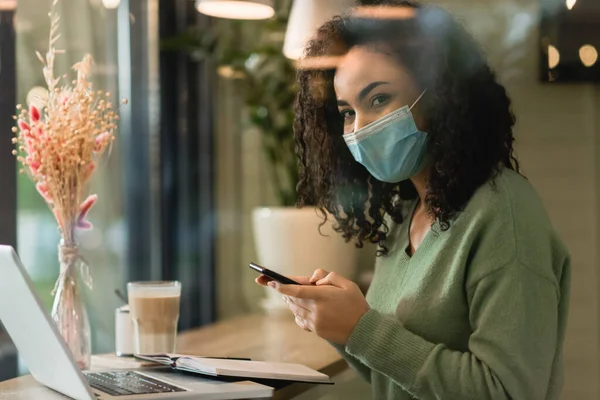 Afrikanerin Mit Medizinischer Maske Hält Smartphone Neben Laptop Und Glas — Stockfoto