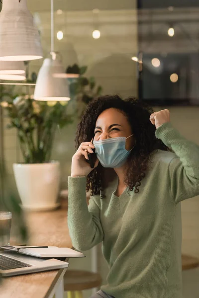 Excited African American Woman Medical Mask Talking Smartphone Laptop Cafe — Stock Photo, Image