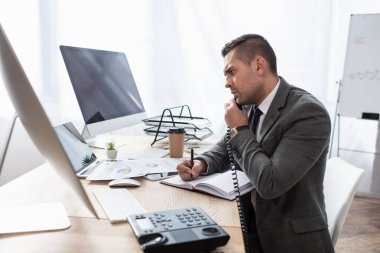 bilgisayar monitörleri ve not defterinin yanında defterde yazarken telefonda konuşan ciddi bir işlemci, bulanık önplan