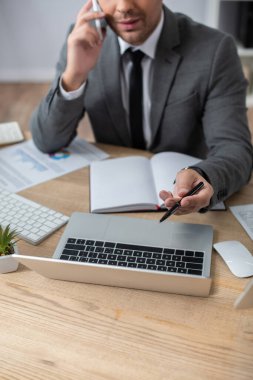 Akıllı telefondaki bulanık arkaplanda konuşurken dizüstü bilgisayarı işaret eden kırpılmış simsar görüntüsü