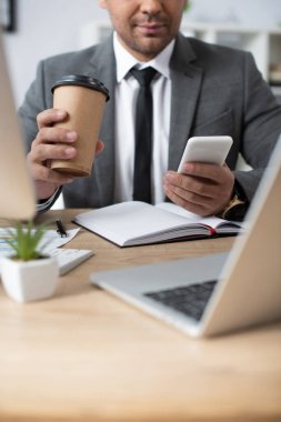 Önplanda bulanık dizüstü bilgisayarın yanında akıllı telefondan mesajlaşırken elinde kahve olan tüccarın kırpılmış görüntüsü