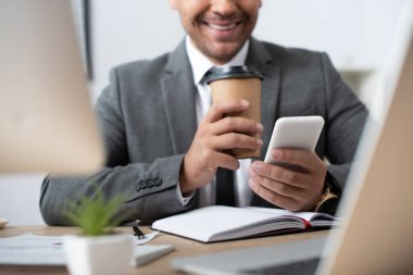 cropped view of smiling businessman chatting on smartphone while holding coffee to go on blurred foreground clipart