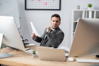 İş yerinde oturup monitöre bakarken elinde kağıt tutan işadamı