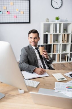 smiling trader holding coffee to go while writing in notebook near computer monitor on blurred foreground clipart