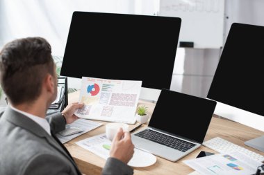 Bilgisayarın yanında bilgi grafikleri ve boş ekranlı monitörler tutan tüccar ön planı bulanıklaştırdı