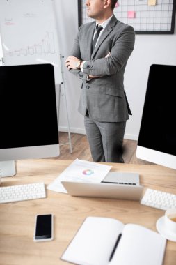 Dizüstü bilgisayarın yanında çapraz kollu duran tüccarın kısmi görüntüsü ve boş ekranlı monitörler