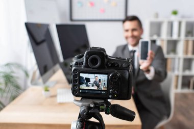 İş adamının yakınındaki dijital kameranın seçici odağı bulanık arkaplanda video yayını sırasında boş ekranlı akıllı telefonu tutuyor