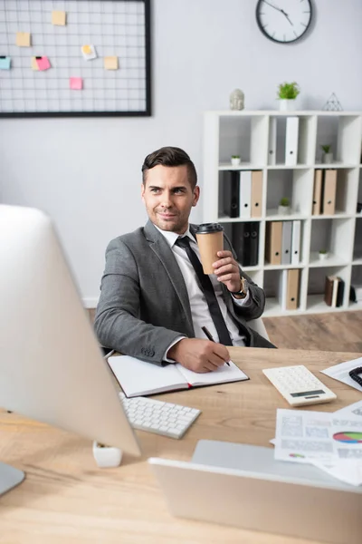 Smiling Trader Holding Coffee While Writing Notebook Computer Monitor Blurred — Stock Photo, Image