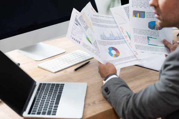 partial view of trader holding infographics near laptop with blank screen, blurred foreground