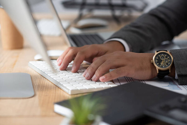 cropped view of trader in wristwatch typing on keyboard on blurred foreground