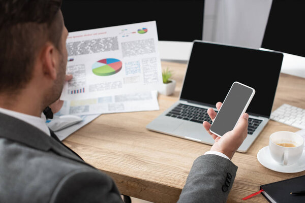 partial view of trader holding smartphone and infographics near laptop and coffee cup, blurred foreground