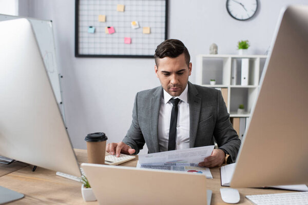 trader looking at paper while sitting near coffee to go and computers on blurred foreground