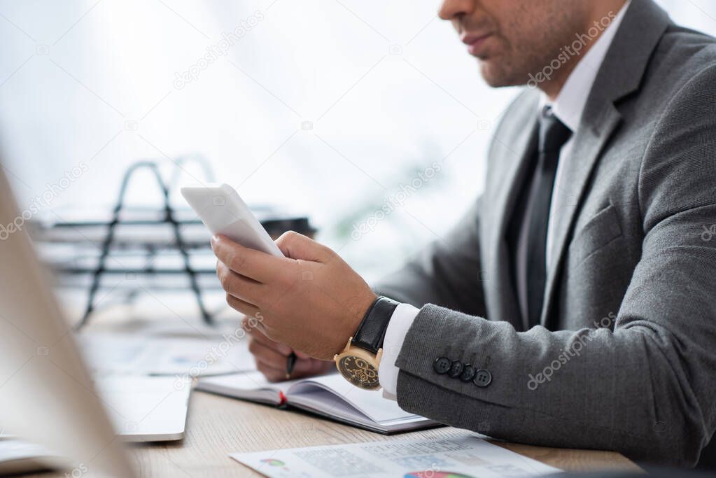 cropped view of businessman messaging on smartphone at workplace on blurred foreground