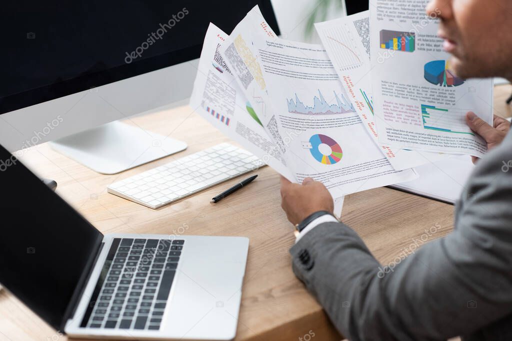 partial view of trader holding infographics near laptop with blank screen, blurred foreground