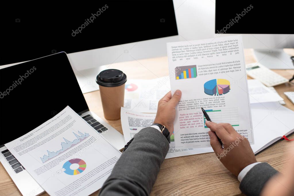 partial view of trader pointing with pen at infographics near laptop and coffee to go