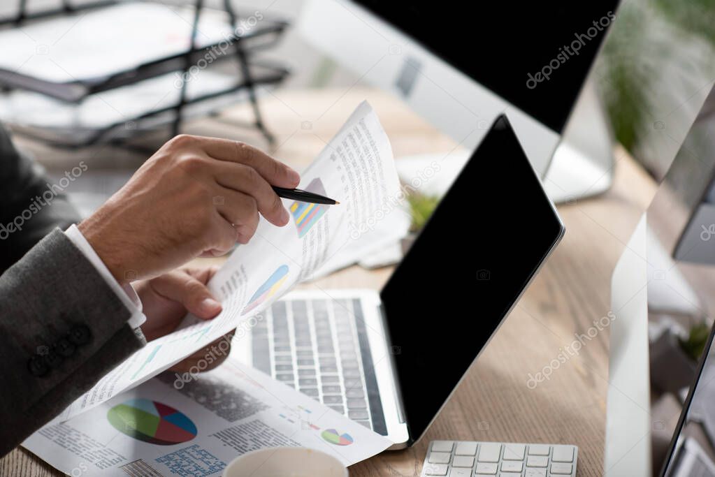 cropped view of trader pointing with pen at infographics near laptop with blank screen