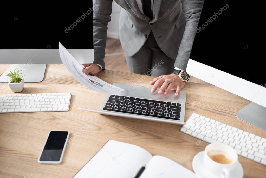 partial view of trader holding infographics near laptop, monitors and smartphone with blank screen