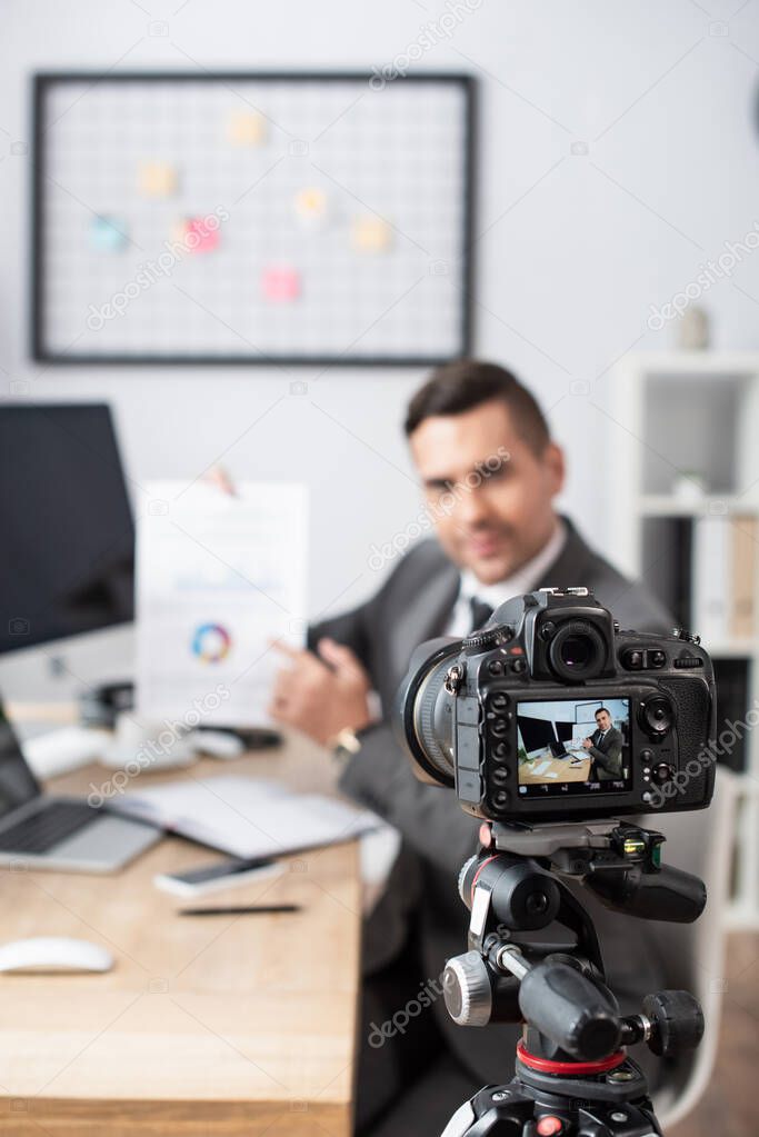 selective focus of digital camera near trader pointing with finger at infographics during online streaming on blurred background