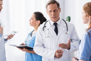 Düşünceli doktor klinikte not defteri ve dijital tabletle çok ırklı meslektaşlarının yanında hemşire ile konuşuyor. 