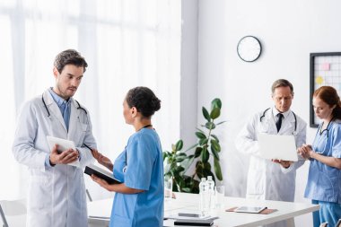 Dijital tabletli ve defterli çok ırklı hastane personeli klinikteki bulanık arka planda laptopla iş arkadaşlarının yanında konuşuyor. 