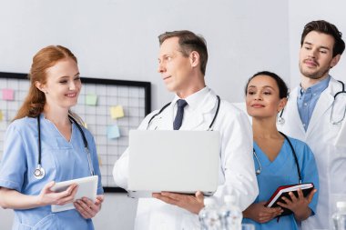 Klinikte çalışırken gülümseyen dijital tabletli ve dizüstü bilgisayarlı çok ırklı hastane personeli 