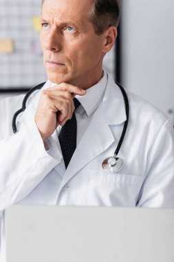 Klinikteki bulanık önplanda dizüstü bilgisayarı tutan düşünceli bir doktorun görüntüsü. 