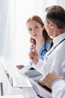 Bilgisayarlı doktor parmak ucunda dijital tabletli hemşire ve bulanık önplanlı meslektaşı gösteriyor. 