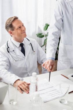 Gülümseyen doktor meslektaşının yanına oturmuş ön plandaki kağıtlara ve su şişelerine işaret ediyor. 