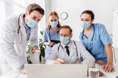 Tıp maskeli çok ırklı doktor ve hemşireler hastanedeki iş yerinde bilgisayarlarını bulanık ön planda kullanıyorlar.