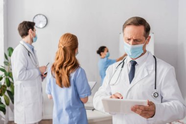 Tıbbi maskeli doktor arka planda bulanık etnik meslektaşlarıyla dijital tablet kullanıyor.