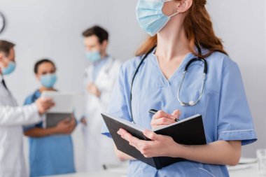 Tıbbi maskeli kızıl hemşire arka plan üzerinde çalışan çok ırklı meslektaşlarıyla birlikte deftere yazı yazıyor.