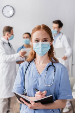 Sağlık maskeli kızıl saçlı hemşirenin arka planda çok kültürlü meslektaşlarıyla kameraya baktığı ön manzara.