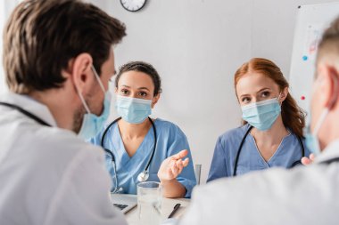Tıp maskeli çok kültürlü hemşireler iş yerinde bulanık ön planda otururken doktorlara bakıyorlar.