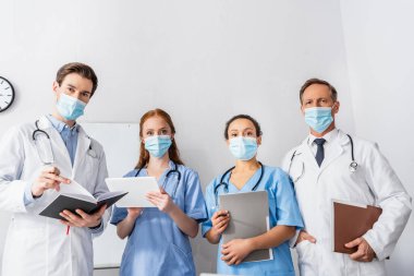 Evrakları ve dijital tabletleri olan çok ırklı hemşire ve doktorlar hastanede yan yana dururken kameraya bakıyorlar.