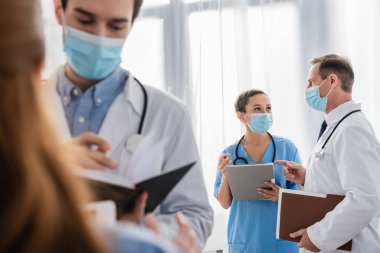 Kağıt klasörlü Afrikalı Amerikalı hemşire ön planda bulanık meslektaşlarıyla doktorla konuşuyor.