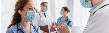 Kızıl saçlı hemşire arka planda, pankartta bulanık çok kültürlü meslektaşlarıyla deftere yazan doktorla konuşuyor.