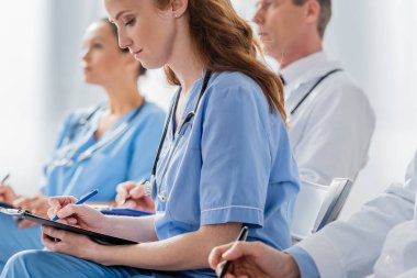 Redhead nurse writing on clipboard while sitting near multicultural colleagues during meeting in hospital on blurred background clipart