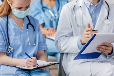 Nurse writing on clipboard while sitting near doctor during meeting with blurred colleagues on background in hospital clipart