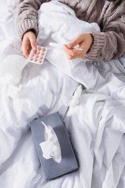 cropped view of sick young woman with tissue, pills and thermometer in bed clipart