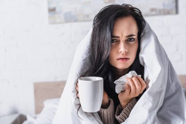 sick young brunette woman with tissue and hot drink in bed clipart