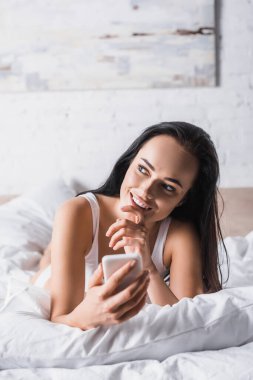 Hayalperest, genç, esmer kadın yatakta akıllı telefon kullanıyor.